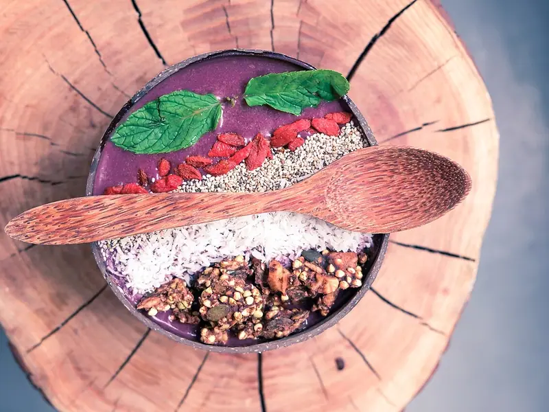 Purple smoothie bowl in a coconut shell topped with goji berries, chia seeds, shredded coconut, granola and mint, with a wooden spoon