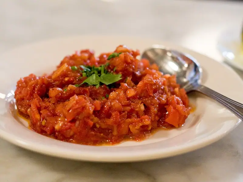 O movilă de salată fin tocată din roșii și ardei, presărată cu pătrunjel, pe o farfurie albă, cu o lingură, pe o masă de marmură.
