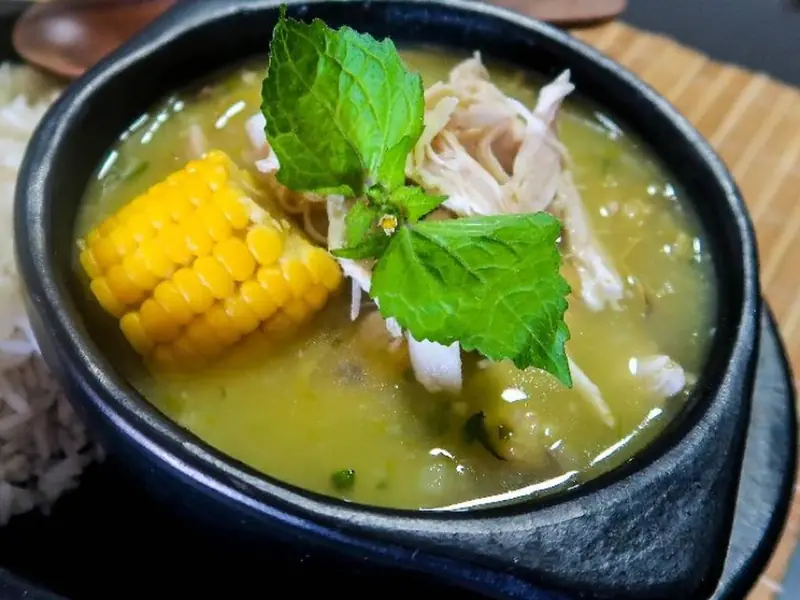 Sort skål med cremet grøngul kyllingesuppe med et stykke majs og en myntelignende urt ved siden af hvide ris og skiver af avocado på en sort tallerken.