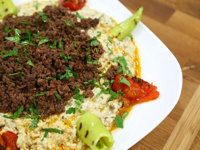 Angebratenes Hackfleisch mit Petersilie auf cremigem Auberginen-Joghurt-Püree, dazu gegrillte grüne Spitzpaprika und geröstete Tomaten auf einer weißen Platte.