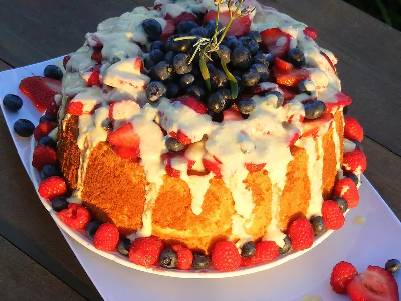 Tall golden tube cake drizzled with white glaze, piled with blueberries and strawberries, ringed with raspberries and topped with lavender.