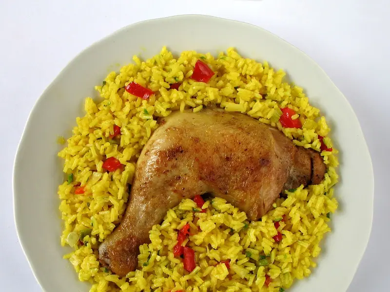 Uma coxa e sobrecoxa de frango assada com pele sobre arroz amarelo vibrante com pimentão vermelho em cubos e cebolinha, em um prato branco.