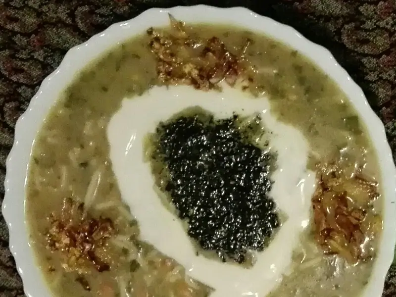 Desenli bir örtü üzerinde, beyaz kaşk kıvrımı, ortasında koyu renkli naneli yağ ve altın rengi kızarmış soğanla süslenmiş bir kase yeşil otlu erişte çorbası.
