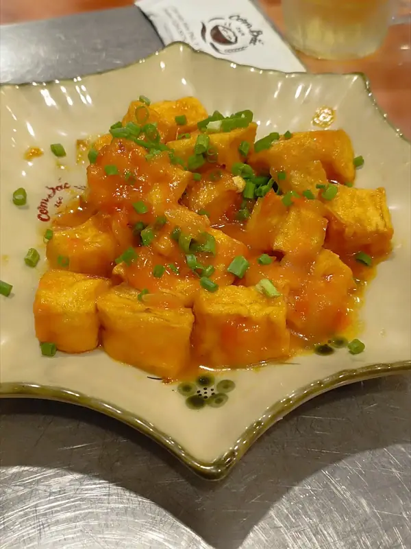 Zlatisté kocky tofu obalené lesklou oranžovou paradajkovou omáčkou a posypané nasekanou jarnou cibuľkou na keramickom tanieri v tvare hviezdy.