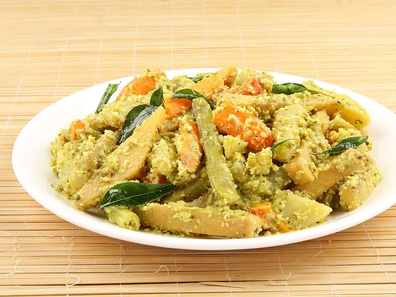 Mixed vegetable batons of carrot, beans and plantain coated in a coarse coconut paste with curry leaves on a white plate on a bamboo mat.