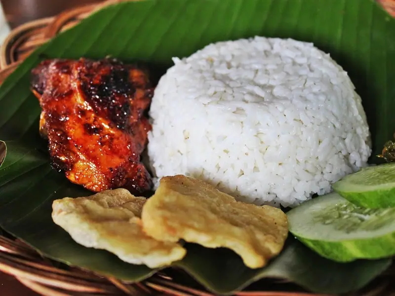 Glazed, charred grilled chicken with a dome of white rice, fried crackers, cucumber slices and chili sambal on a banana leaf in a basket.