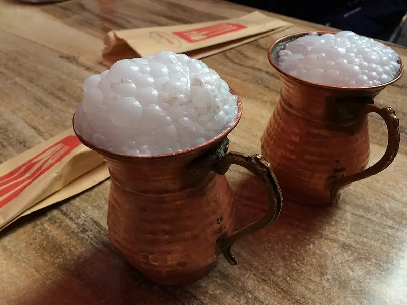 To hamrede kobberkrus med ayran toppet med højt hvidt skum på et træbord ved siden af papirindpakning fra sugerør.