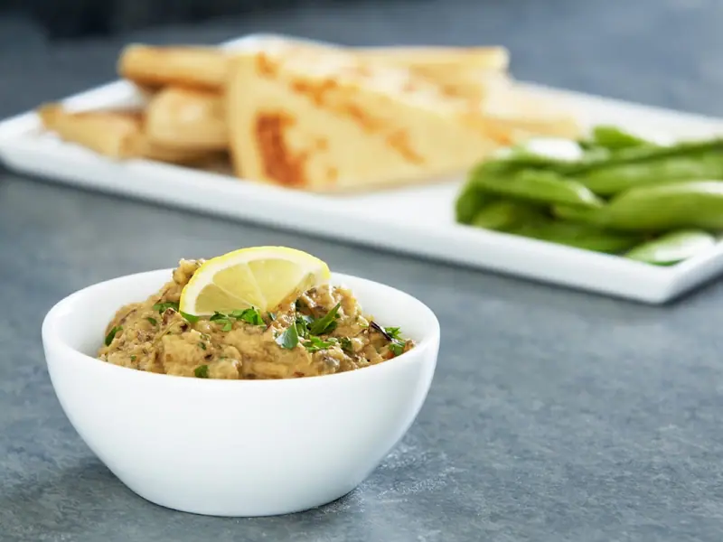 En lille hvid skål med grov auberginedip, toppet med en citronbåd og hakket persille, med grillet pitabrød og sukkerærter bagved.