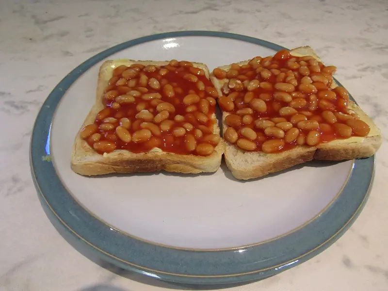 To skiver smurt, ristet hvidt brød med baked beans i tomatsauce på en hvid tallerken med blågrå kant.