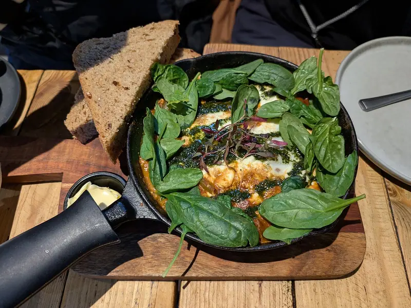Æg bagt i en krydret tomatsauce i en lille støbejernspande, toppet med grøn krydderurteolie, frisk spinat og mikrogrønt, serveret med ristet brød.