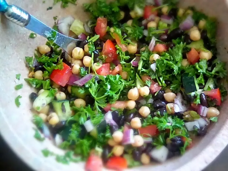 Kichererbsen-Salat mit schwarzen Bohnen, gewürfelten Tomaten, Gurke, roter Zwiebel und reichlich gehackter Petersilie in einer beigefarbenen Schüssel mit Löffel.