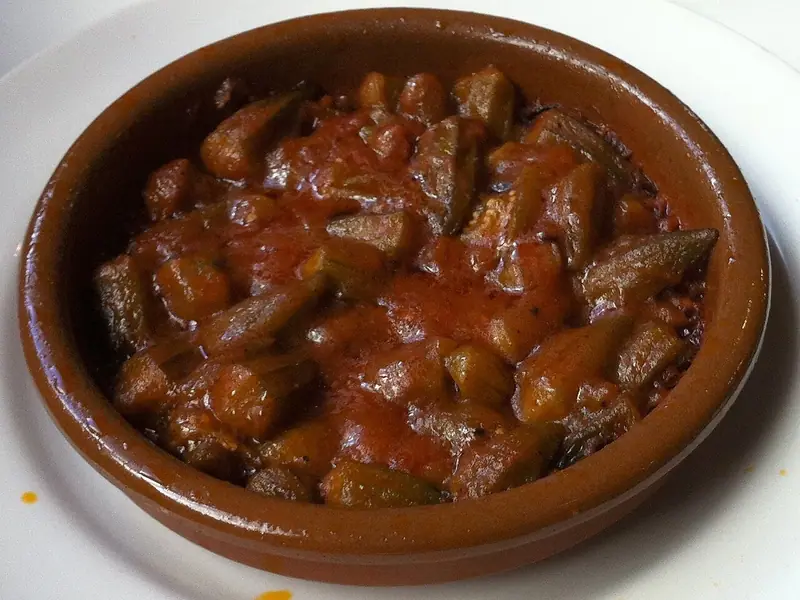 Okra pieces in a thick red tomato sauce served in a small glazed terracotta dish set on a white plate.