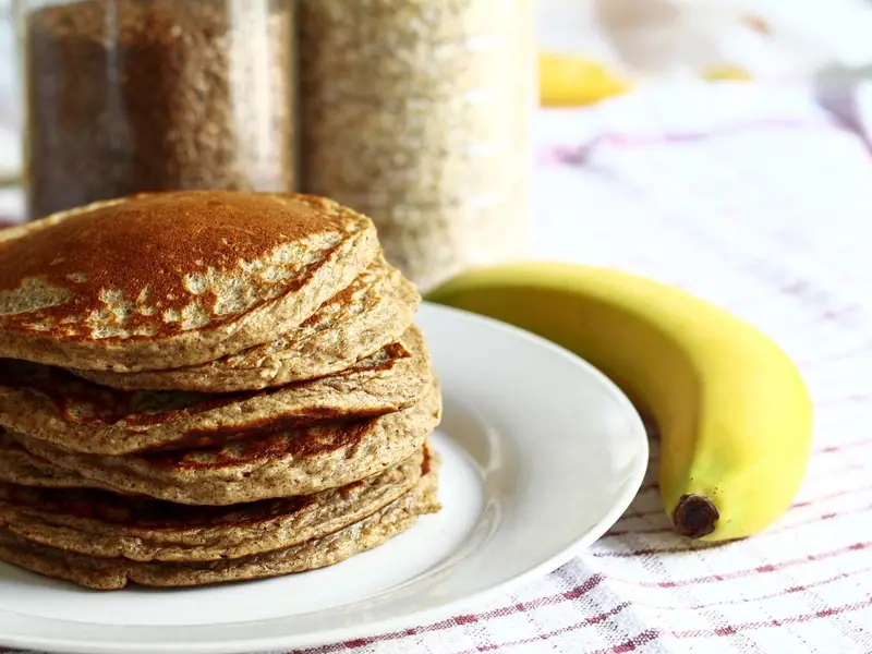 Una pila alta de tortitas de avena doradas en un plato blanco junto a un plátano amarillo, con tarros de avena al fondo sobre un mantel de cuadros.