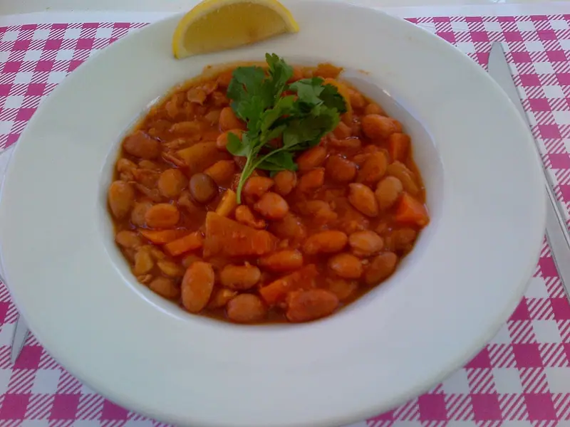 Borlotti bean stew with carrot and potato in red tomato sauce in a white rimmed bowl, topped with parsley, lemon wedge, pink check cloth