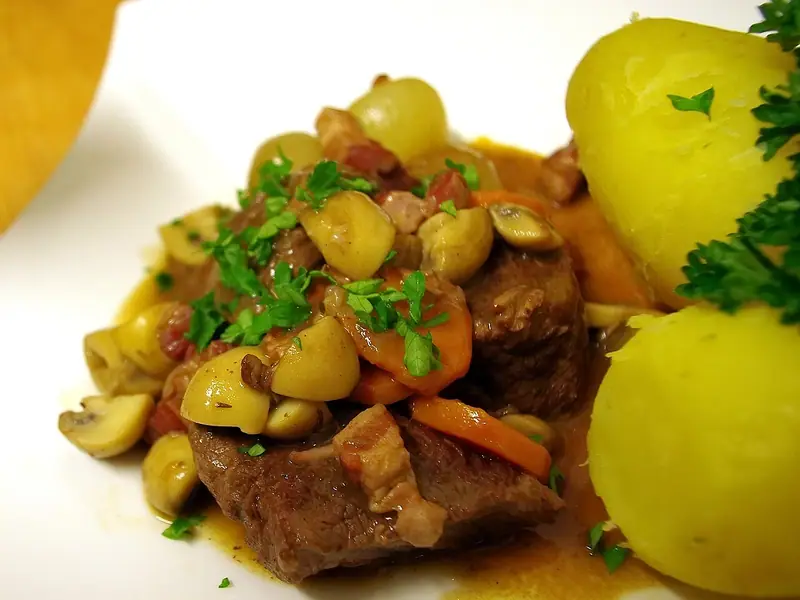 Oksegryde med champignoner, perleløg, gulerødder og bacon i en blank sauce, drysset med persille ved siden af kogte kartofler på en hvid tallerken
