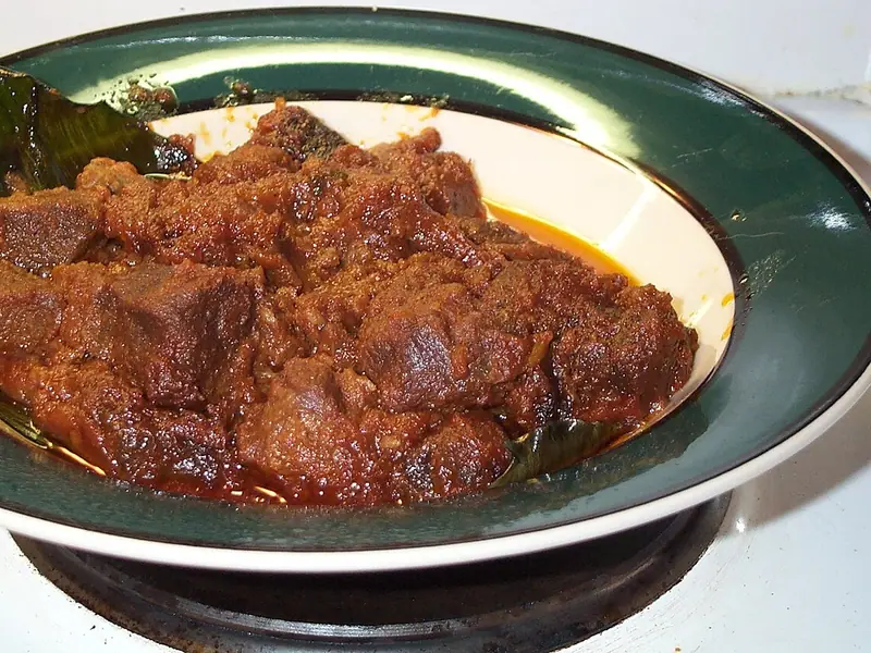 Dark, saucy chunks of beef rendang piled in a green-rimmed bowl lined with a banana leaf, with orange oil pooling at the edge.