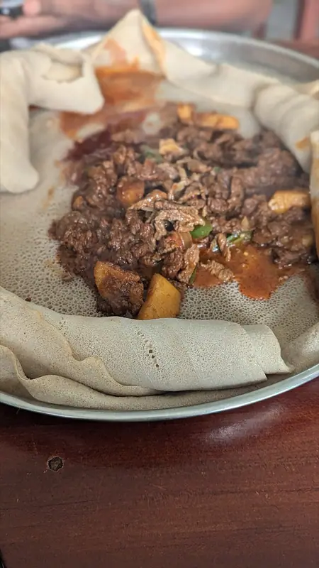 Saftig gryderet med hakket oksekød, kartoffelstykker og grøn chili på en rund injera, med rullede injera på en metalbakke.