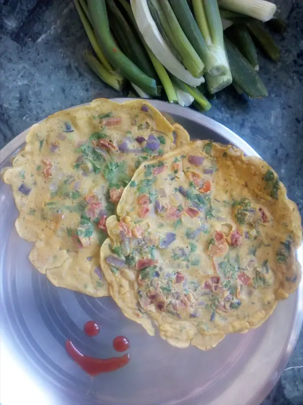 Zwei goldbraune Kichererbsenmehl-Pfannkuchen mit Zwiebel-, Tomaten- und Korianderstückchen auf einem Stahlteller, dazu ein Ketchup-Smiley und Frühlingszwiebeln