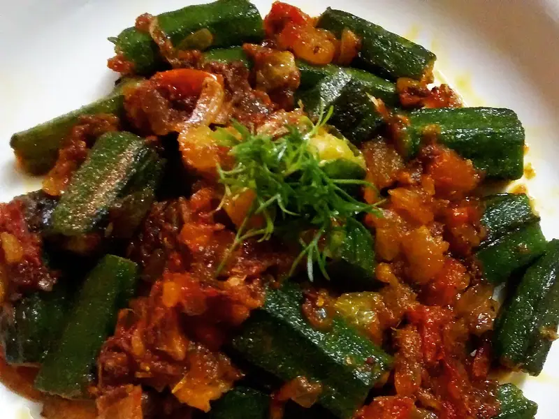 Bucăți de bame amestecate într-o masala roșiatică-maronie de ceapă și roșii, pe o farfurie albă, ornate cu o crenguță de mărar proaspăt sau frunze de fenicul.