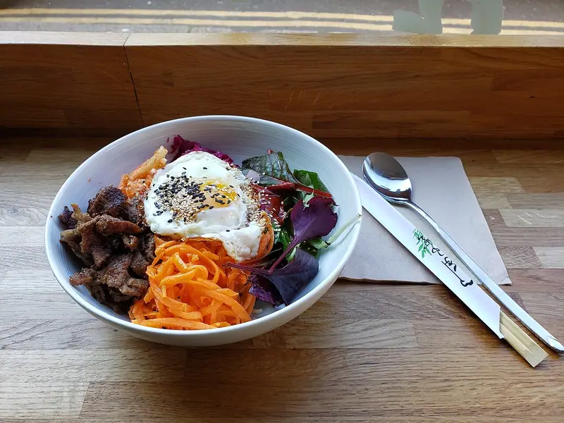 White bowl of bibimbap with browned beef, shredded carrot, kimchi, red cabbage, mixed leaves and a fried egg with sesame seeds