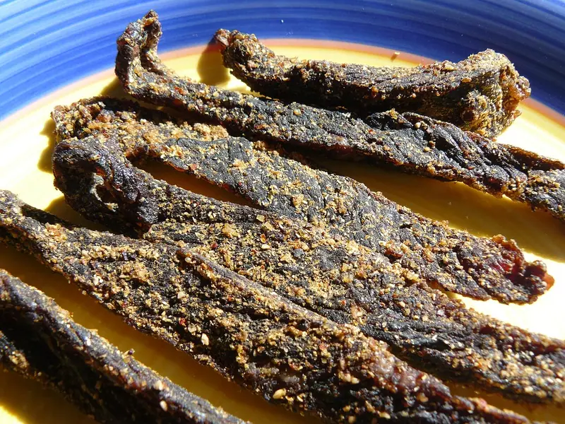 Dark, chewy strips of dried beef coated in crushed coriander and pepper, laid on a yellow plate with a blue rim in bright sunlight.