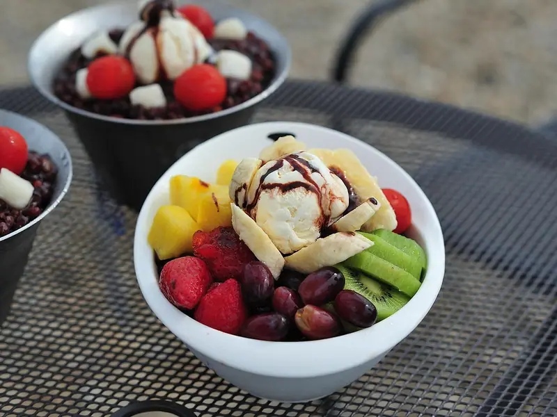 White bowl of mango, strawberries, kiwi, red grapes and banana around a scoop of ice cream with chocolate drizzle, on a metal table