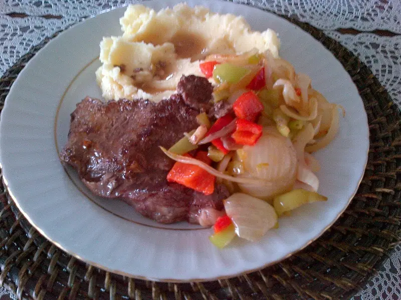 Seared steak on a white gold-rimmed plate with mashed potatoes and gravy, sautéed onions and red and green peppers, on a woven placemat