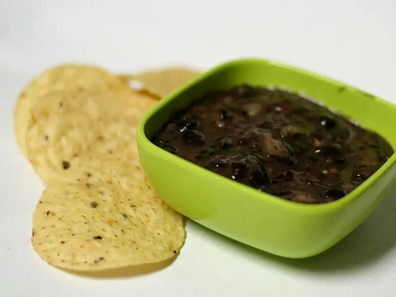 Chunky dark black bean dip with bits of onion and herbs in a square lime-green bowl beside round tortilla chips on a white surface.
