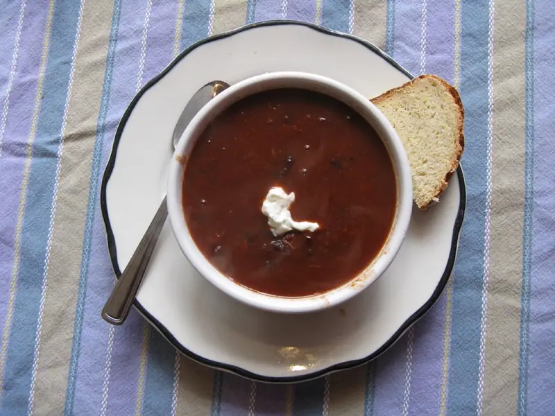 Tigela de sopa de feijão-preto escura e cremosa com uma colherada de creme azedo, sobre um prato branco com uma colher e uma fatia de pão, em um pano listrado.