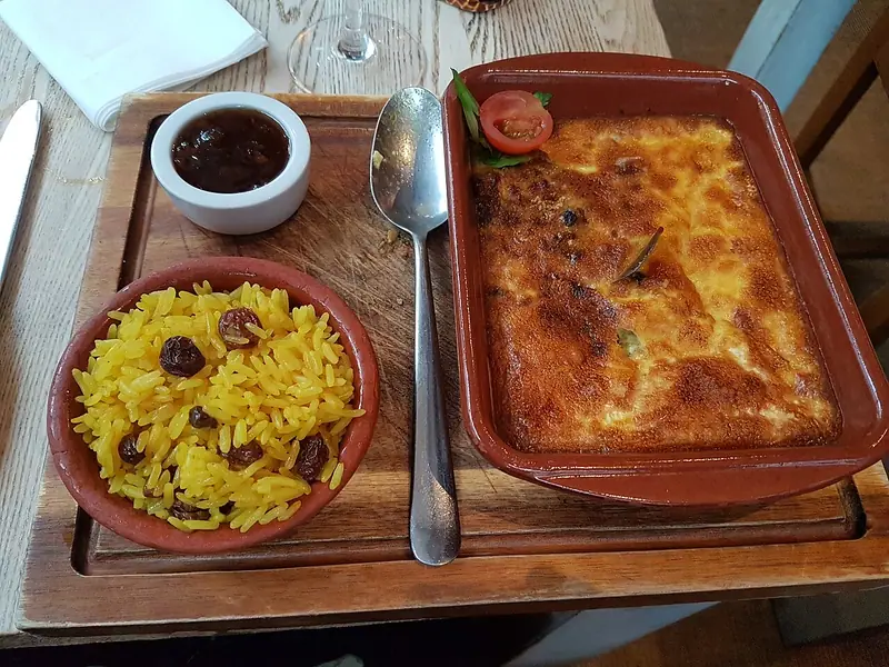 Ahşap bir tahta üzerinde, kuru üzümlü sarı pilav kasesi ve küçük bir kase chutney'nin yanında toprak bir kapta altın renginde fırınlanmış bobotie