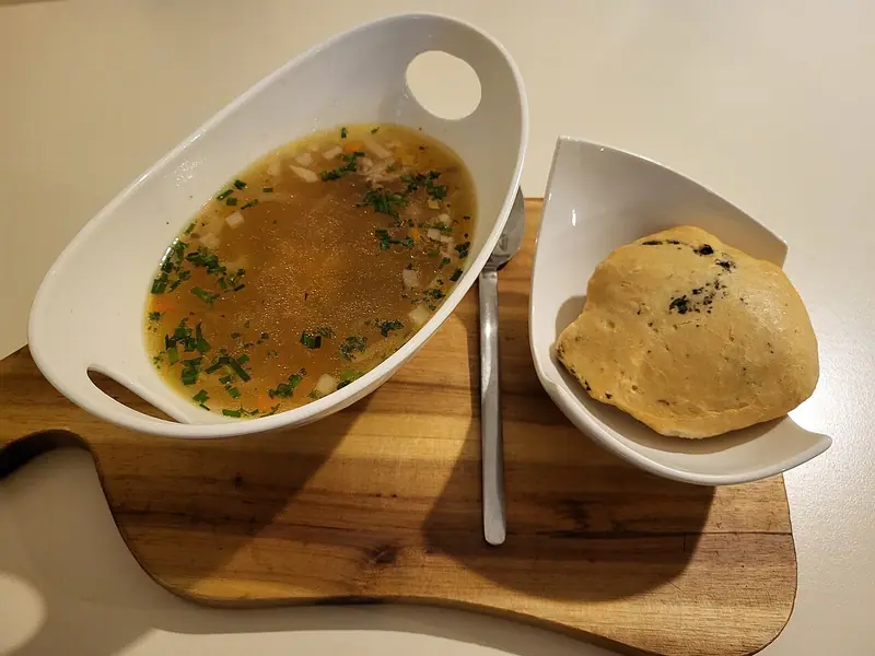Clear golden soup with diced vegetables and chopped chives in a white handled bowl, beside a crusty bread roll on a wooden board.