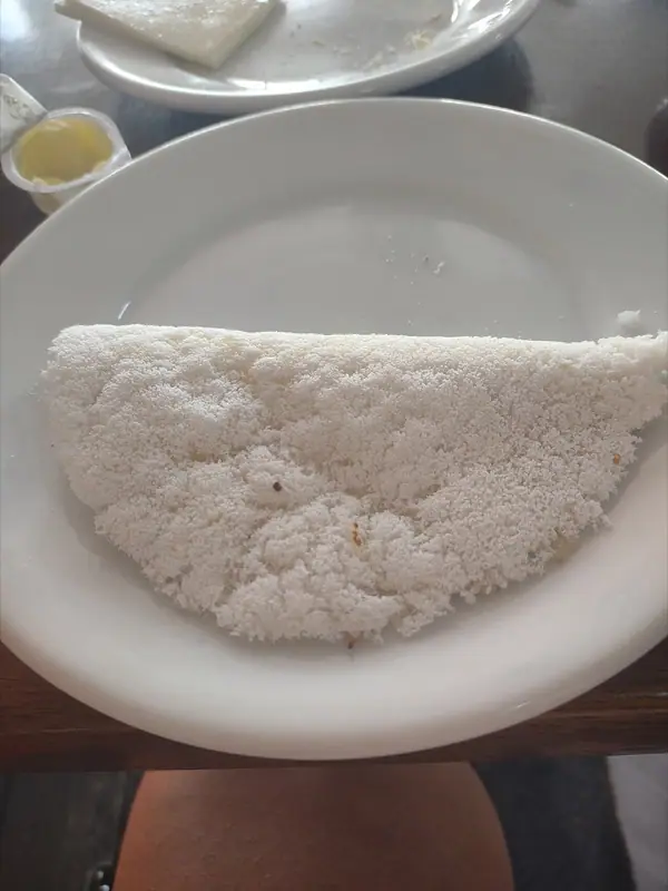 A folded white tapioca crepe on a white plate, with a small cup of butter and another plate behind it on a wooden table.