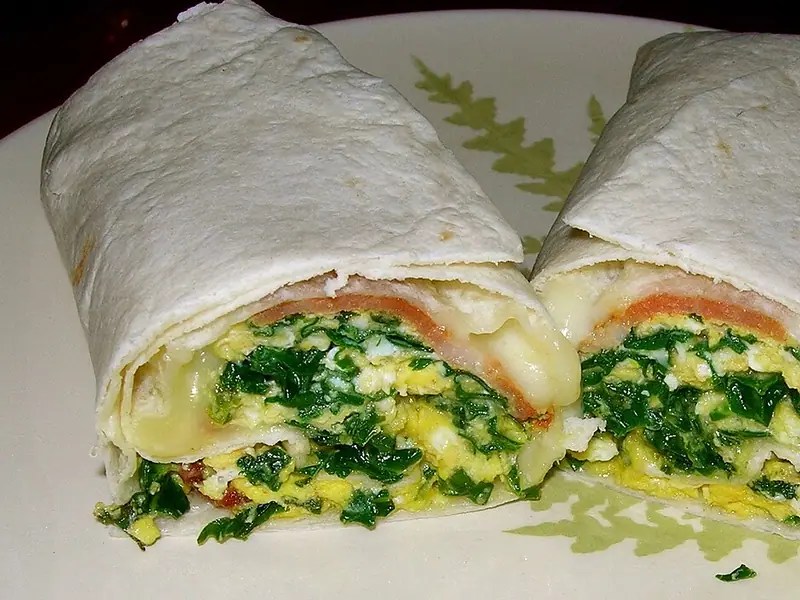 Ein halbierter Tortilla-Wrap mit Rührei, Spinat, geschmolzenem Käse und einer dünnen orangefarbenen Schicht auf einem Teller mit Blattmuster.