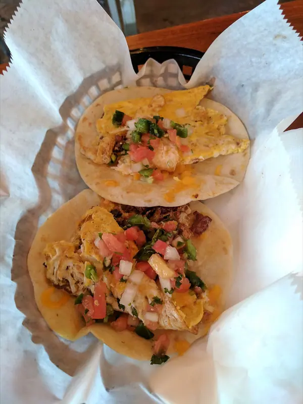 Two flour tortilla breakfast tacos with scrambled egg, chopped meat, melted cheese and pico de gallo in a paper-lined basket.