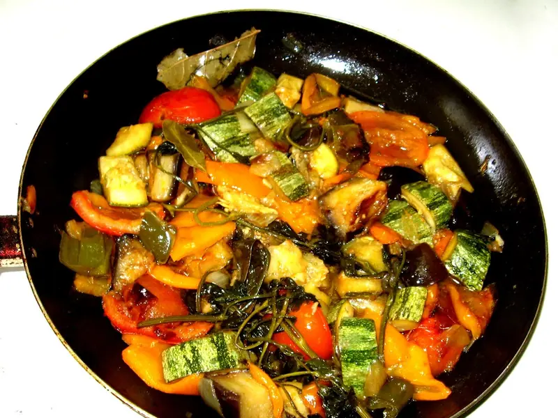Stewed zucchini, eggplant, orange and red peppers and tomato with bay leaves and wilted greens in a black frying pan.