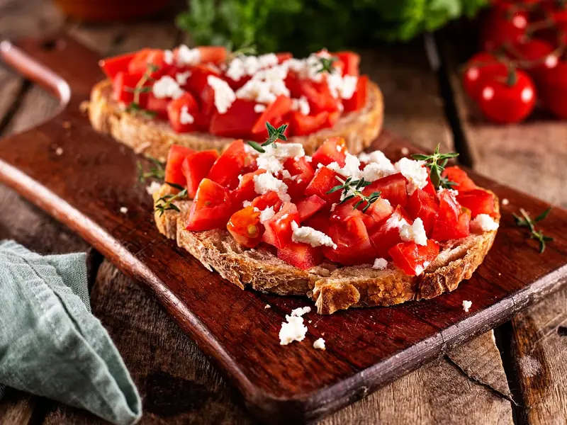 To skiver ristet rustikt brød med tomater i tern, smuldret hvid ost og timian på et træbræt med cherrytomater i baggrunden.