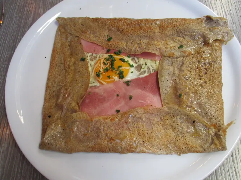 Galette quadrada de trigo-sarraceno em um prato branco, com as bordas dobradas sobre o presunto, o queijo derretido e uma gema brilhante, polvilhada com cebolinha picada.
