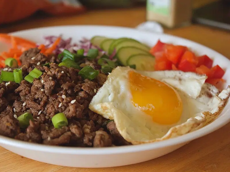 Bowl of browned ground beef with spring onions and sesame seeds, a fried egg, carrots, red cabbage, cucumber and red pepper.