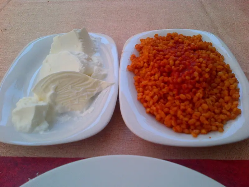 Zwei weiße Teller nebeneinander auf einer rosa Tischdecke: auf dem einen weicher weißer Käse oder Butter, auf dem anderen orangeroter Bulgur-Pilaw mit Tomaten.