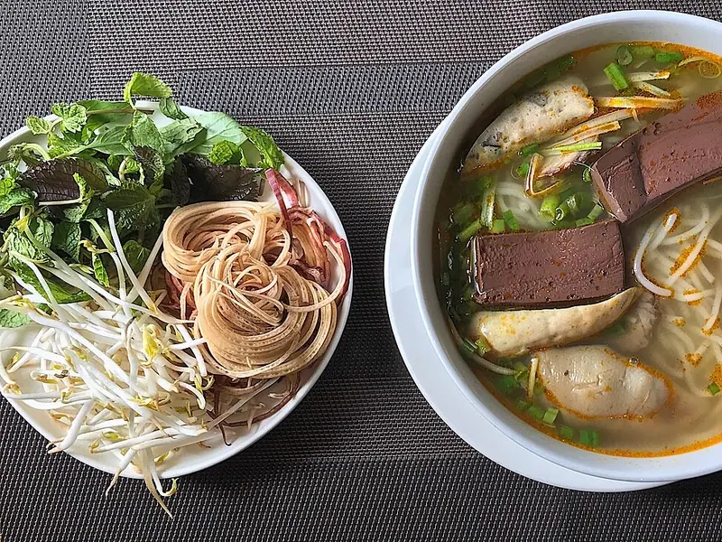 Bowl of bún bò Huế with round noodles, blood cubes, pork sausage and spring onions, beside a plate of herbs, sprouts and banana blossom