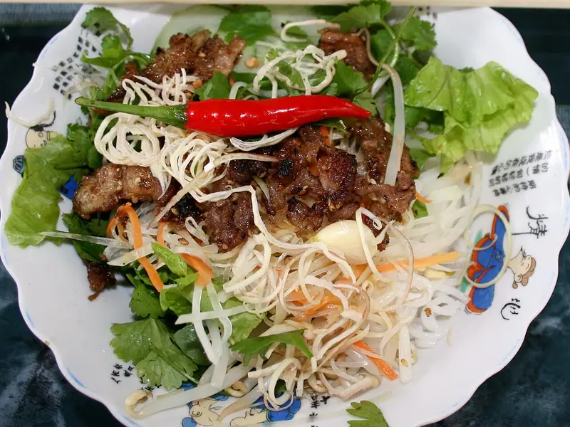 Grillet svinekjøtt med stekeskorpe over risnudler med bønnespirer, gulrot, salat, koriander og en hel rød chili på en tallerken med spisepinner