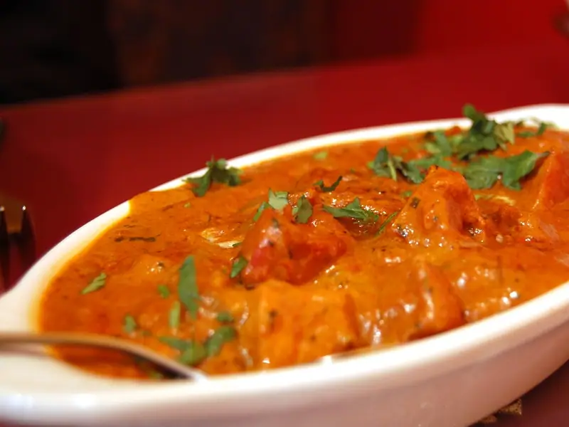 Kremowe pomarańczowe curry pomidorowe z kawałkami kurczaka, posypane posiekaną kolendrą, w białym owalnym naczyniu z łyżką na czerwonym stole