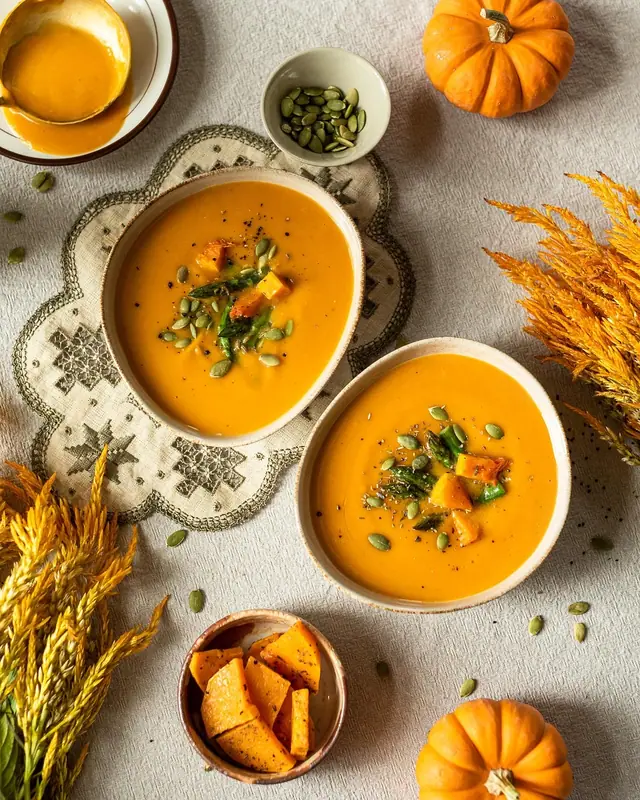 To skåle med glat, orange squashsuppe toppet med ovnbagte squashtern, asparges og græskarkerner, med små græskar ved siden af