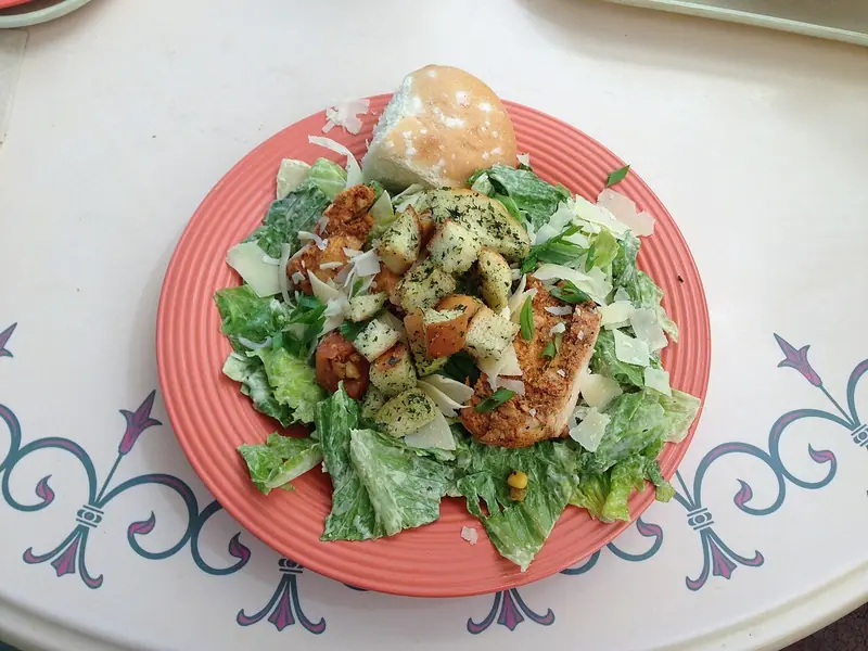 Cæsarsalat på en koralfarvet tallerken med romainesalat vendt i dressing, krydderurtecroutoner, krydret kylling, høvlet parmesan, forårsløg og en bolle