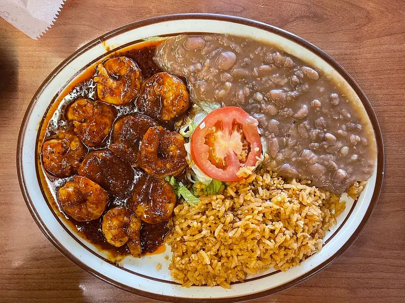 Shrimp in dark red chile sauce on an oval plate with refried pinto beans, Mexican red rice, lettuce and a tomato slice.