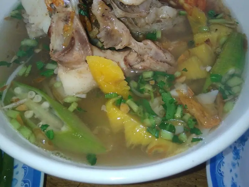 Tigela branca com sopa azedinha de peixe, com pedaços de peixe com espinha, abacaxi, tomate, quiabo, echalota frita e ervas picadas em caldo claro.