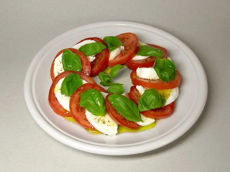 Fatias de tomate e muçarela intercaladas em círculo num prato branco, cobertas com folhas de manjericão, azeite e pimenta-do-reino.