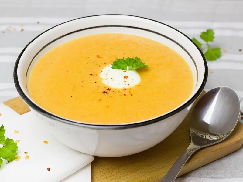 Smooth orange soup in a white bowl topped with a white dollop, a green herb sprig and chilli flakes, with a spoon on a wooden board.