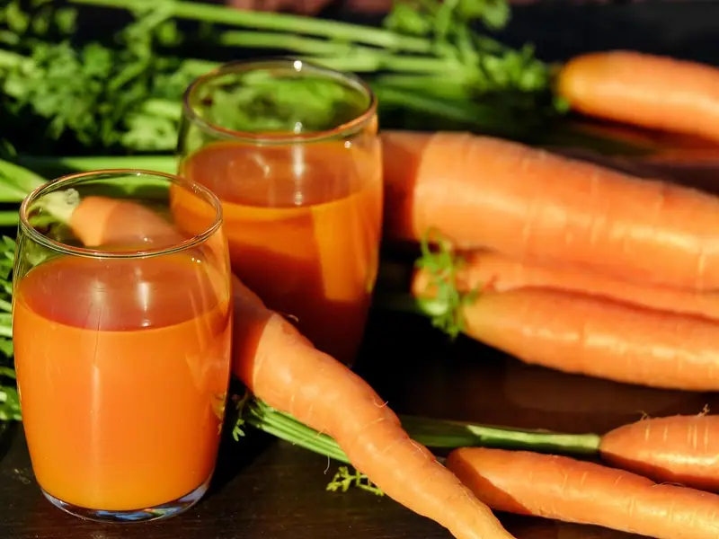 Two glasses of bright orange carrot juice on a dark wooden table beside fresh whole carrots with their green leafy tops.
