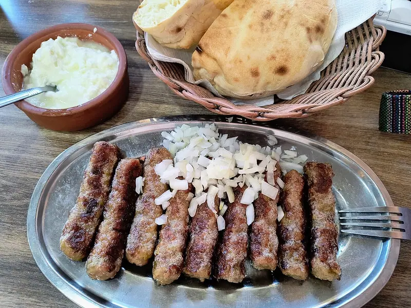Zece mici sârbești (ćevapi) la grătar, presărați cu ceapă crudă tocată, pe un platou metalic, alături de un coș cu lipii pufoase și un bol cu cremă de brânză albă.